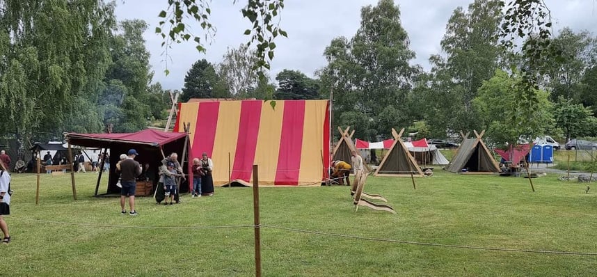 A Viking reenactment village with colorful tents, wooden poles, and participants in period clothing gathered around—recreating daily life in a historical camp setting