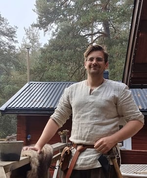A man dressed in Viking-style linen clothing stands confidently on a wooden porch with forest surroundings—part of a historical reenactment.