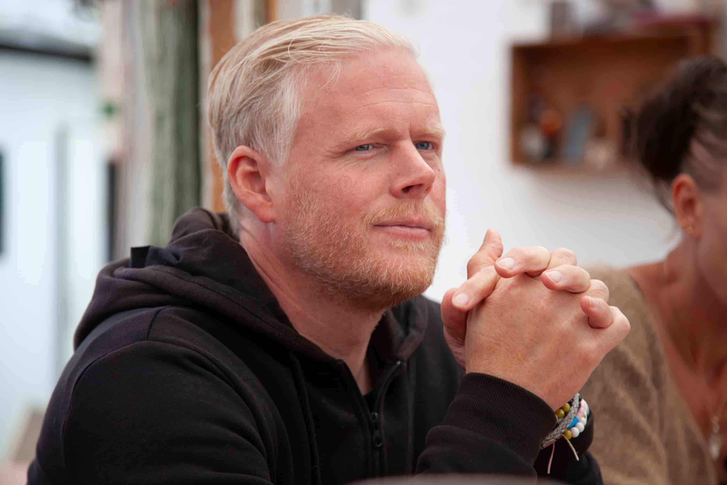 A man with blond hair and a beard looking thoughtfully to the side. He is wearing a black hoodie and has his hands clasped