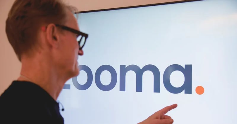 The CEO of Zooma points at the company logo displayed on a large screen.
