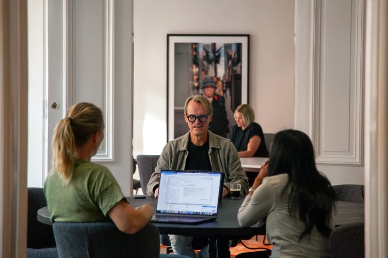 En grupp människor sitter runt ett bord i ett ljust mötesrum på ett kontor och samarbetar över en bärbar dator, med andra som arbetar i bakgrunden och inramade konstverk på väggen.