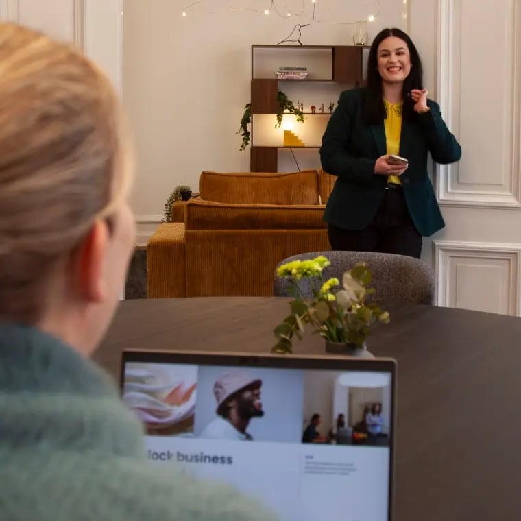 Woman presenting in a meeting room while another person watches from a table with a laptop.