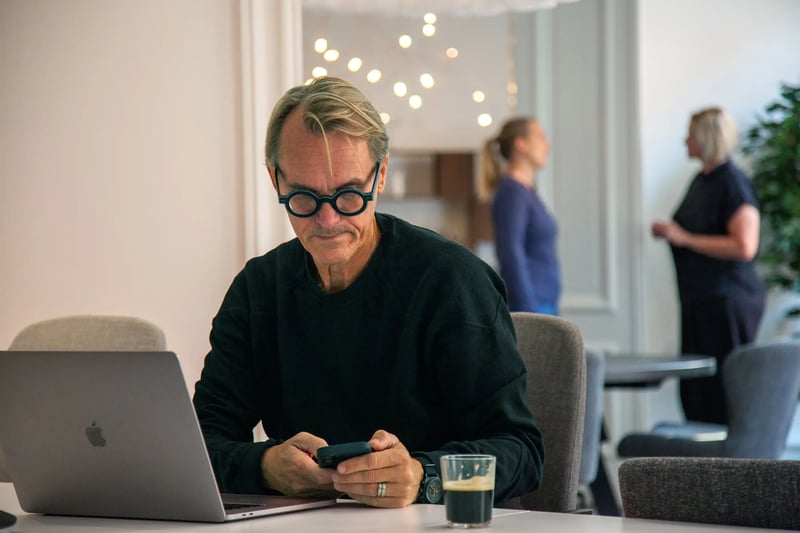 Man with round glasses using his phone beside a laptop, with coffee on the table