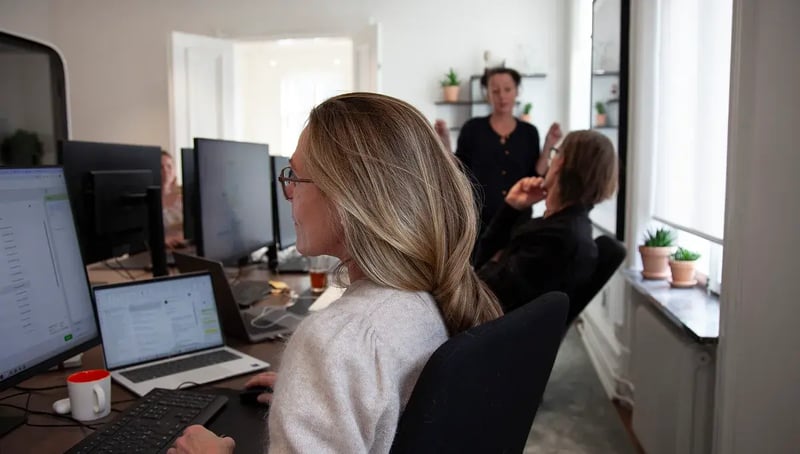 Office workspace with people working at computers and discussing in the background.