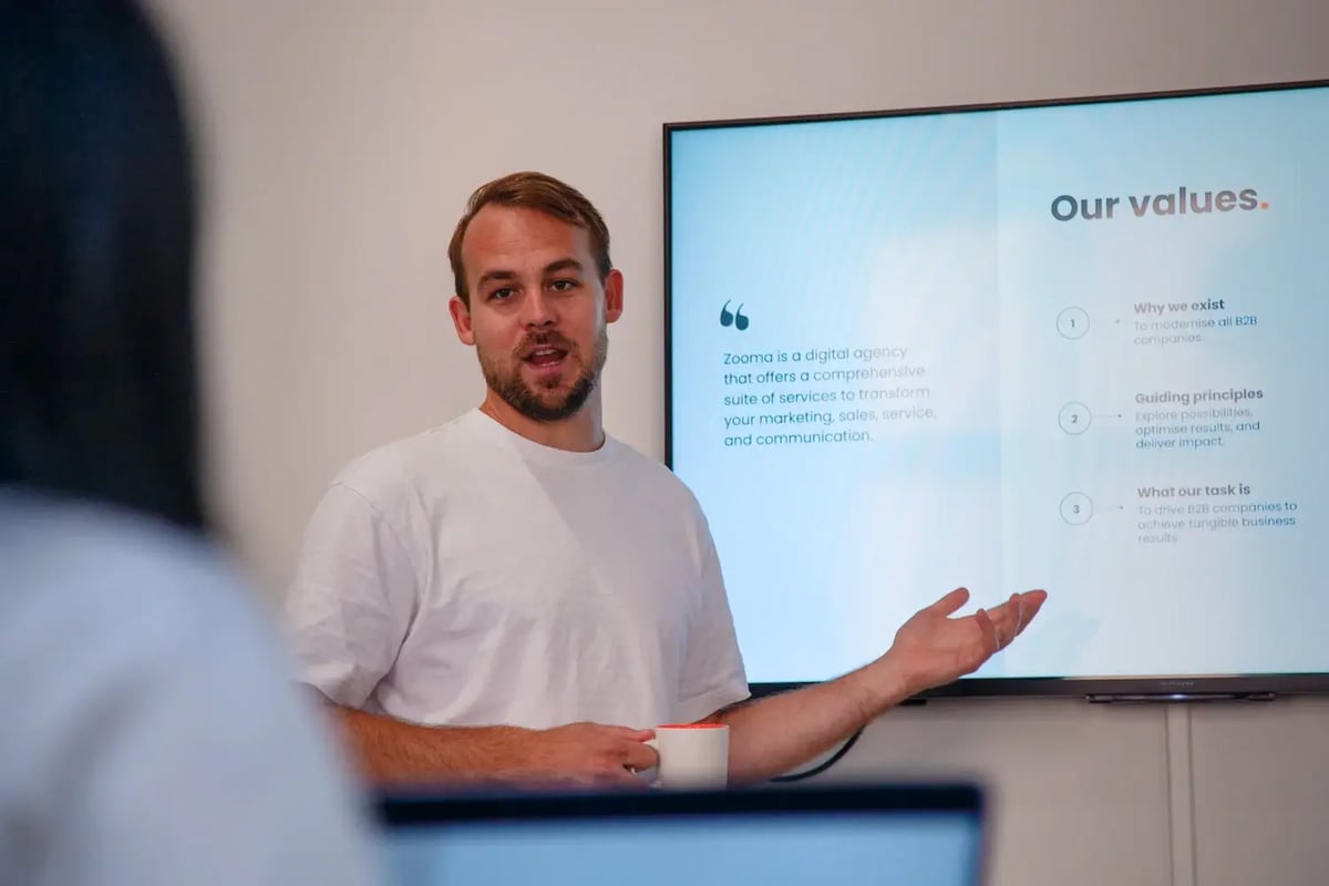 Person som står framför en presentationsskärm under ett möte och gestikulerar samtidigt som han håller i en kopp