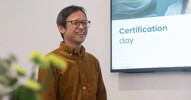 Smiling Zooma colleague standing beside a screen displaying “Certification day” during a learning session.
