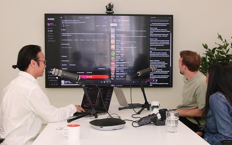 Three people sitting around a table in a modern office, recording a the tech hub podcast or video session with microphones and laptops- A large screen displays a content creation tool, and recording equipment is visible on the table