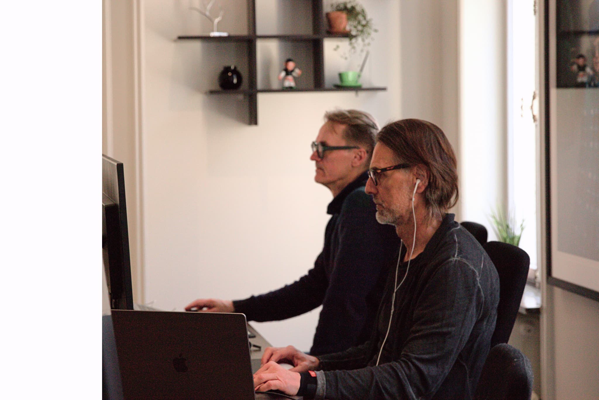 Two colleagues in an office sit side by side watching a computer screen; one wears headphones and listens to music