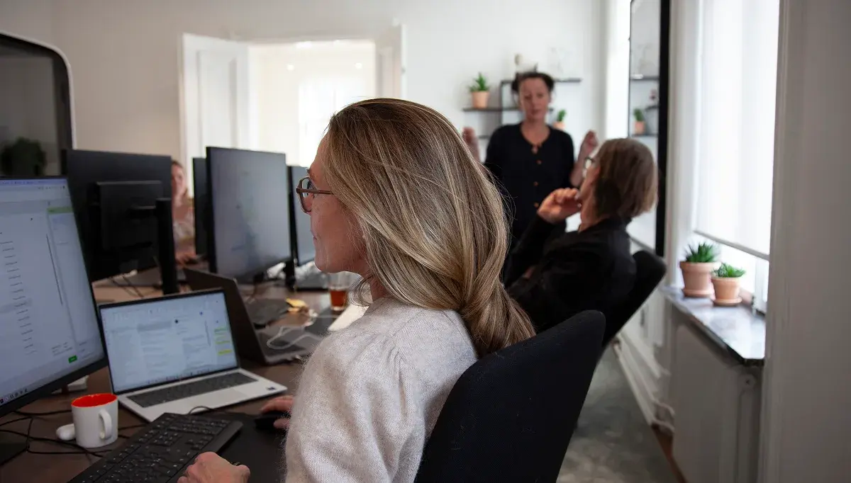 Office workspace with people working at computers and discussing in the background
